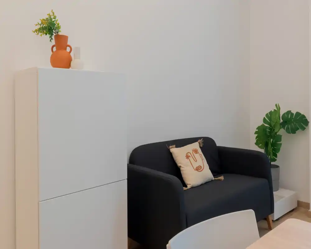 Angolo living moderno e accogliente con un divano scuro compatto, cuscino decorativo e pianta in vaso; pareti neutre e contenitori minimal creano uno spazio calmo e contemporaneo.