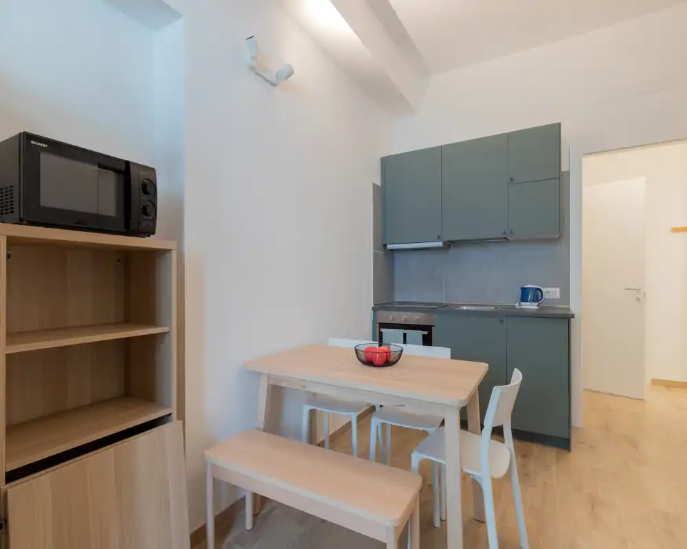 Cucina a vista compatta con mobili verde salvia, piccolo forno e bollitore, tavolo in legno chiaro con panca: ambiente pulito, luminoso e funzionale per un soggiorno accogliente.