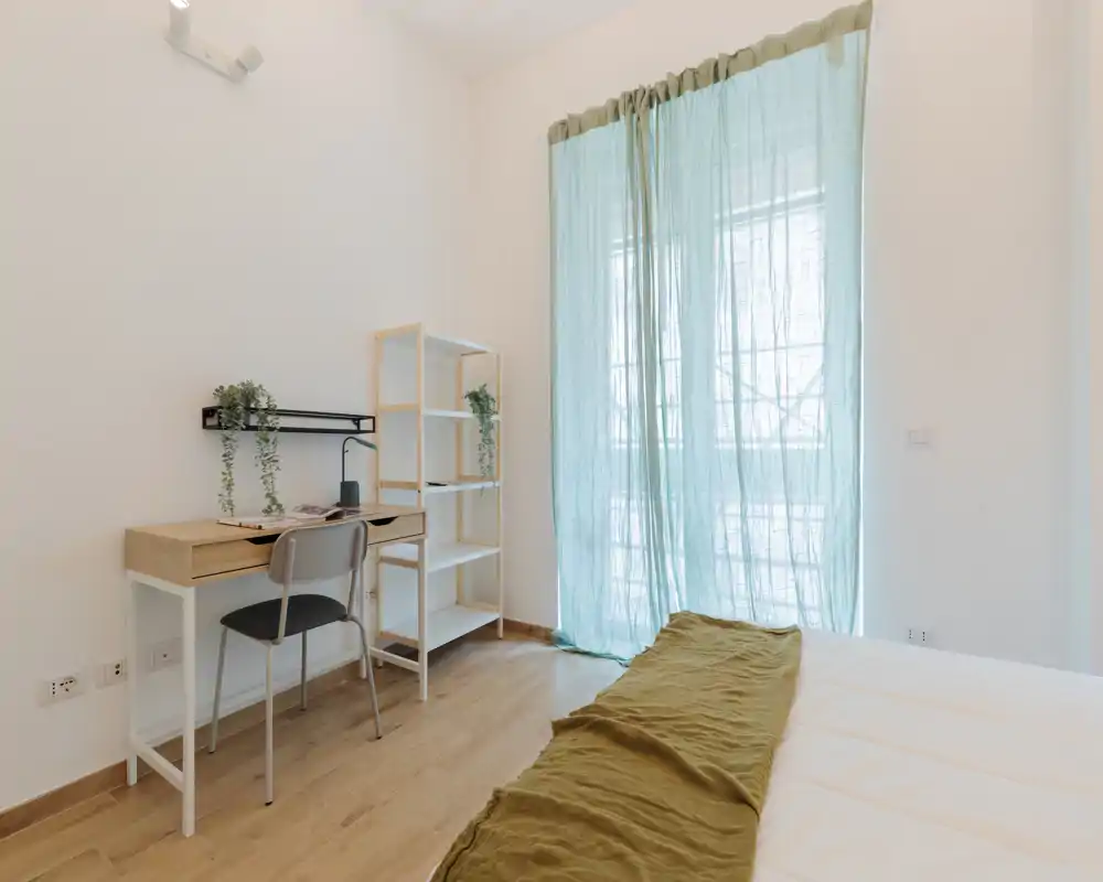 Chambre lumineuse et minimaliste avec lit visible, sol en bois clair, petit bureau et étagères près d'une grande fenêtre avec rideaux légers bleu-vert.