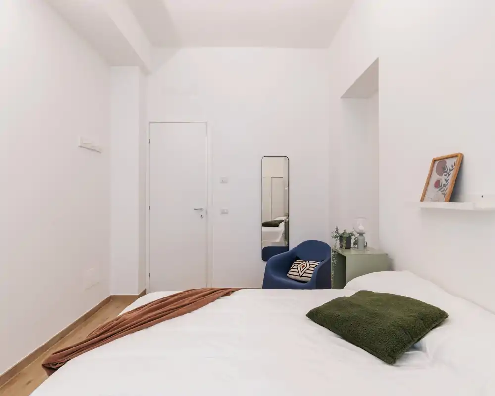 Camera da letto luminosa e minimal con un grande letto in primo piano, tessili accoglienti, una poltrona blu e decorazioni semplici per un'atmosfera tranquilla.