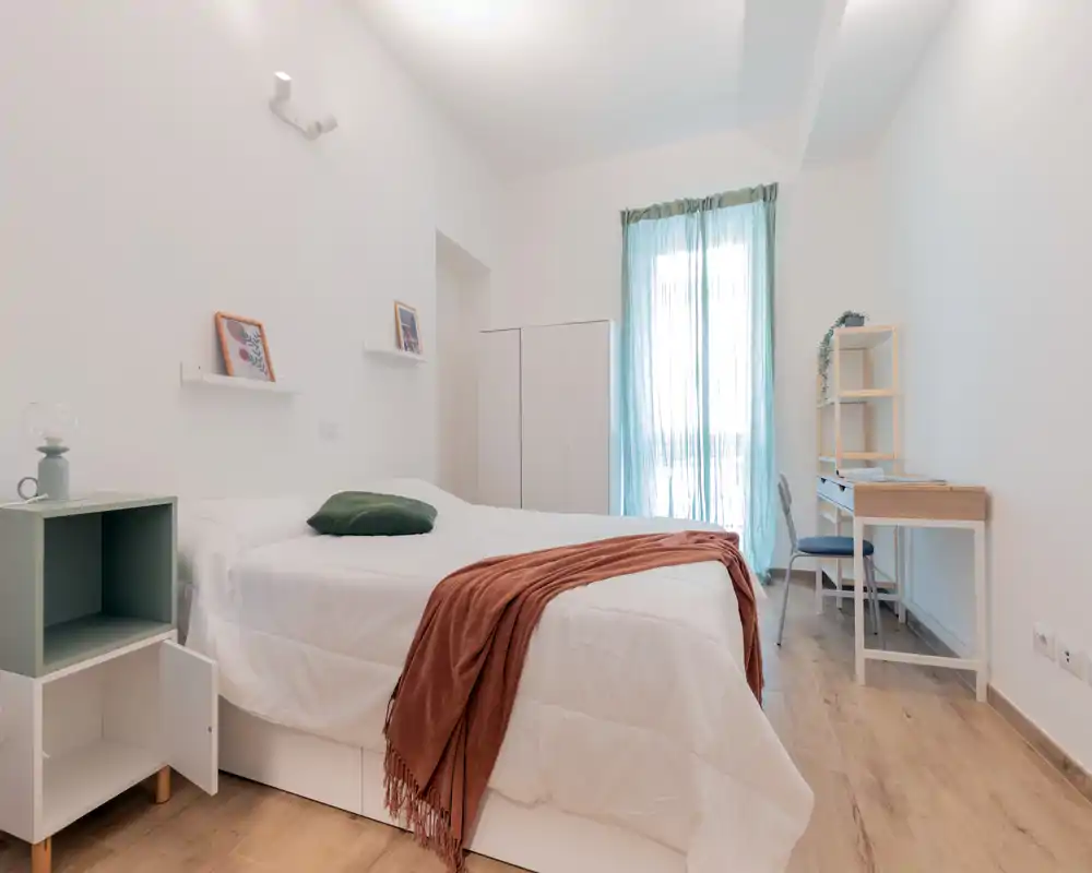 Camera da letto luminosa e ben allestita con un grande letto, biancheria morbida e un plaid accogliente; la luce naturale entra dalla tenda semitrasparente donando aria e luce.