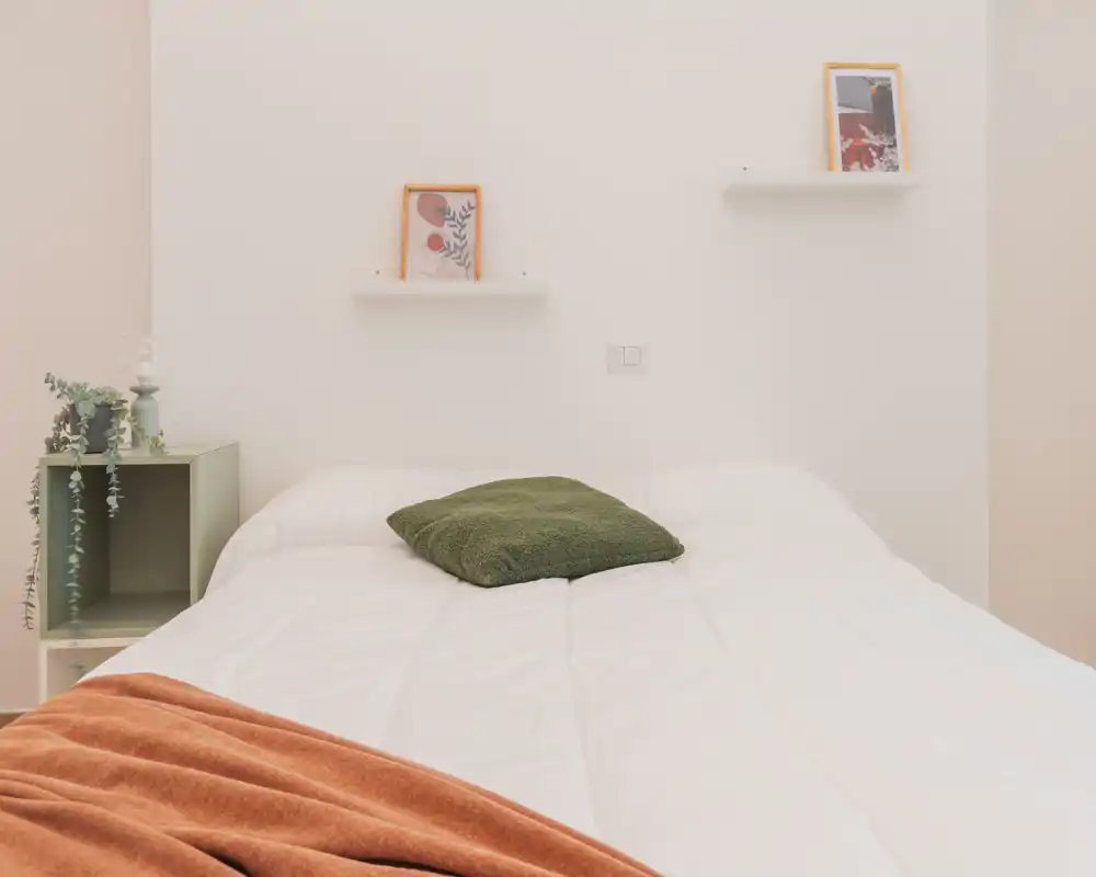 Camera da letto luminosa e minimal con un grande letto con biancheria bianca, un cuscino verde e un plaid color ruggine; mensole semplici e un piccolo comodino a cubo creano un aspetto moderno e tranquillo.