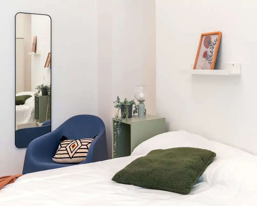 Angolo di camera luminoso e moderno con letto ben rifatto, cuscino verde in tessuto, poltrona blu d'accento e dettagli minimal sul comodino come pianta e lampada.