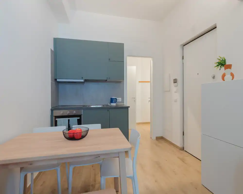 Angolo cottura moderno e compatto con pensili verdi, forno integrato e un tavolo in legno per quattro persone — luminoso, dallo stile semplice e pronto all'uso.