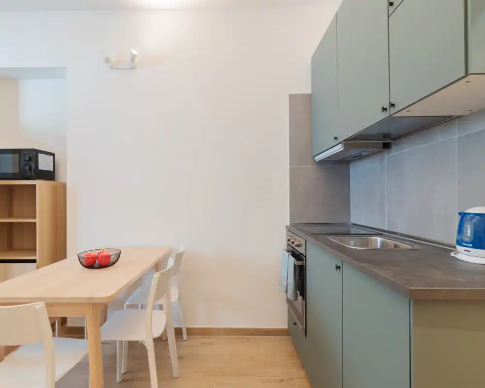 Cucina compatta e moderna con pensili verde tenue, forno incassato e lavello in acciaio. Un tavolo in legno con sedie e una ciotola di frutta decorativa creano un'area pranzo-cucina ordinata e funzionale.