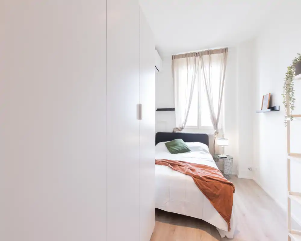 Dormitorio luminoso y minimalista con cama doble, suelo de madera clara y luz natural desde la ventana. Decoración neutra con cojín verde y manta terracota que añaden calidez.