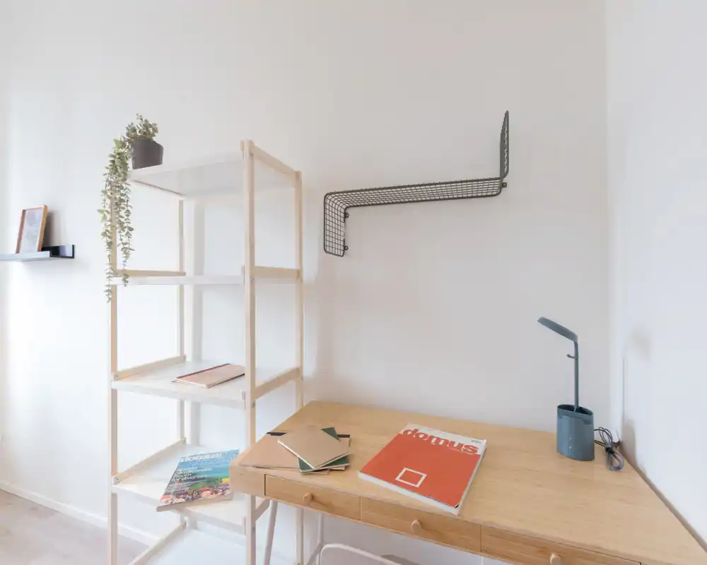 Espacio de trabajo minimalista y luminoso con escritorio de madera, lámpara de sobremesa y estantería abierta; ideal para teletrabajo o estudio en un rincón tranquilo.