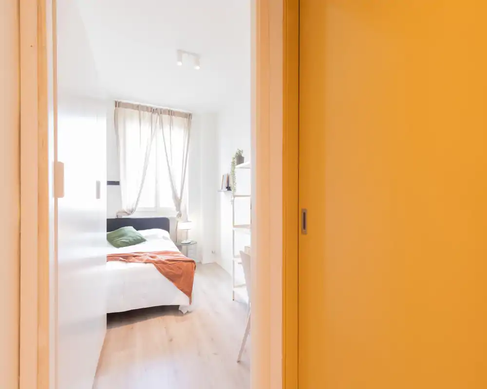 Dormitorio luminoso y compacto con cama doble vestida en blanco, cojín verde y manta naranja. La luz natural de la ventana realza el suelo de madera clara y estanterías minimalistas.