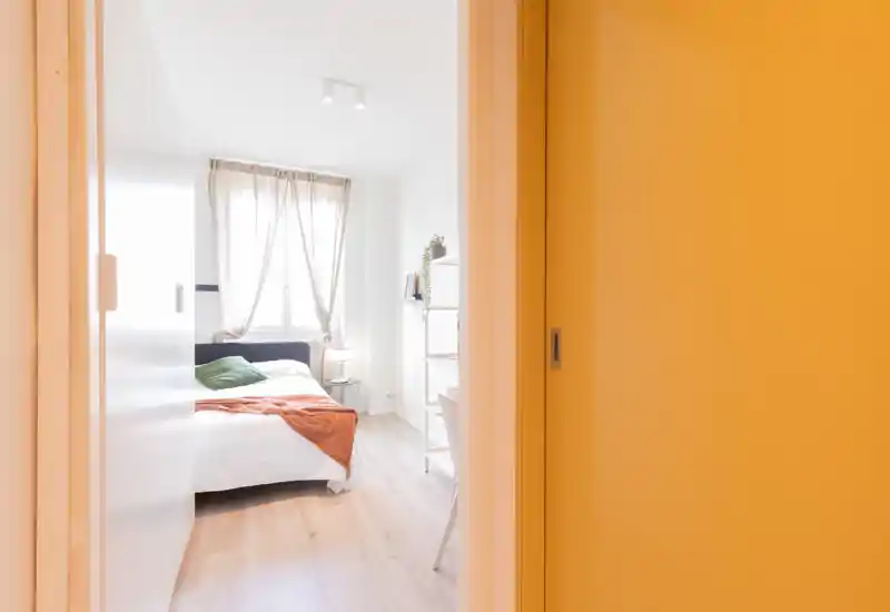 Dormitorio luminoso y compacto con cama matrimonial, suelo de madera clara y acentos cálidos. Cortinas translúcidas que filtran la luz natural; mesita de noche y estantería para almacenamiento.