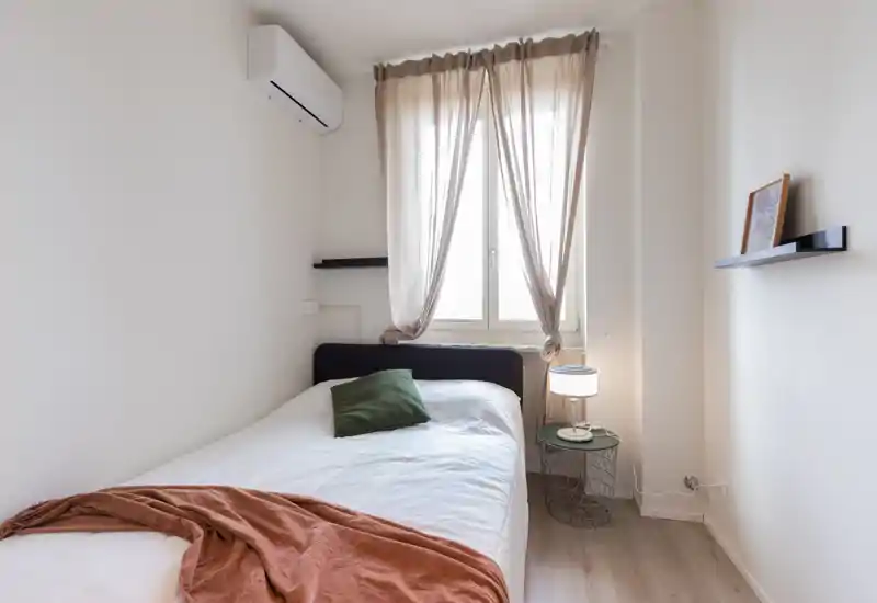 Dormitorio compacto y luminoso con cama individual bien hecha, luz natural filtrada por cortinas finas y decoración moderna y minimalista con lámpara de mesita y pequeña repisa.