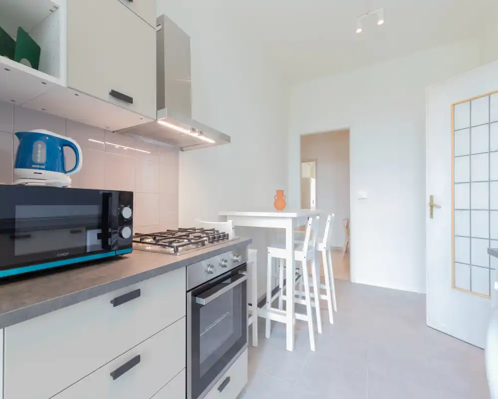 Cocina moderna y limpia con placa de gas, horno empotrado, microondas y una pequeña mesa alta con taburetes; paredes blancas y decoración minimalista que aportan sensación de amplitud.