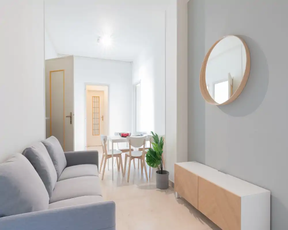 Zona de estar moderna y luminosa con un sofá gris cómodo, un mueble bajo minimalista y un espejo redondo en la pared; una pequeña mesa de comedor al fondo crea una sensación de espacio abierto y aireado.