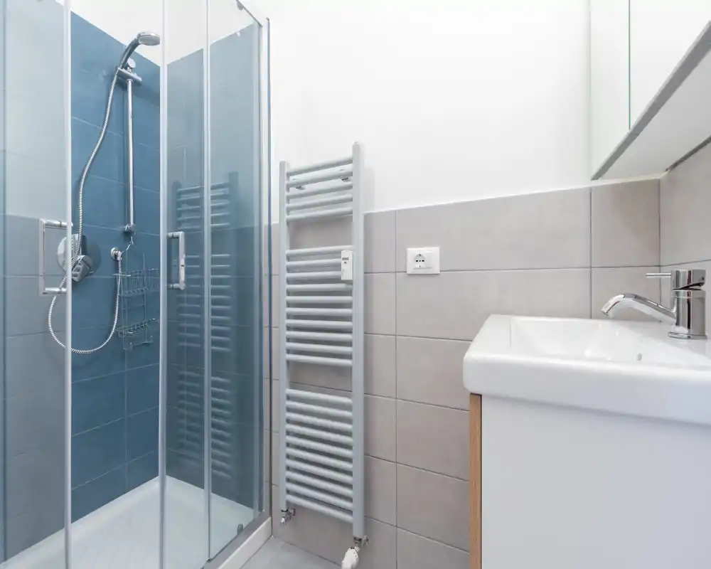 Baño moderno y luminoso con ducha acristalada, toallero calefactado mural y lavabo compacto con grifo cromado — limpio, minimalista y funcional.