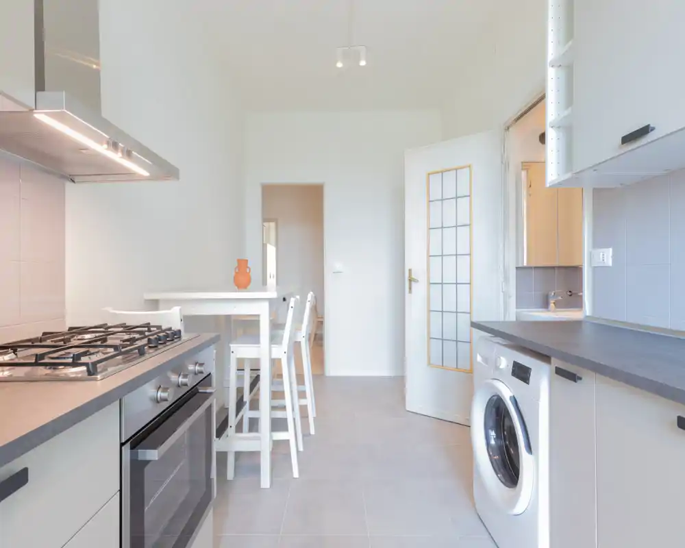 Cocina moderna y luminosa con cocina de gas, horno empotrado, lavadora y una pequeña mesa alta — limpia, minimalista y bien iluminada.