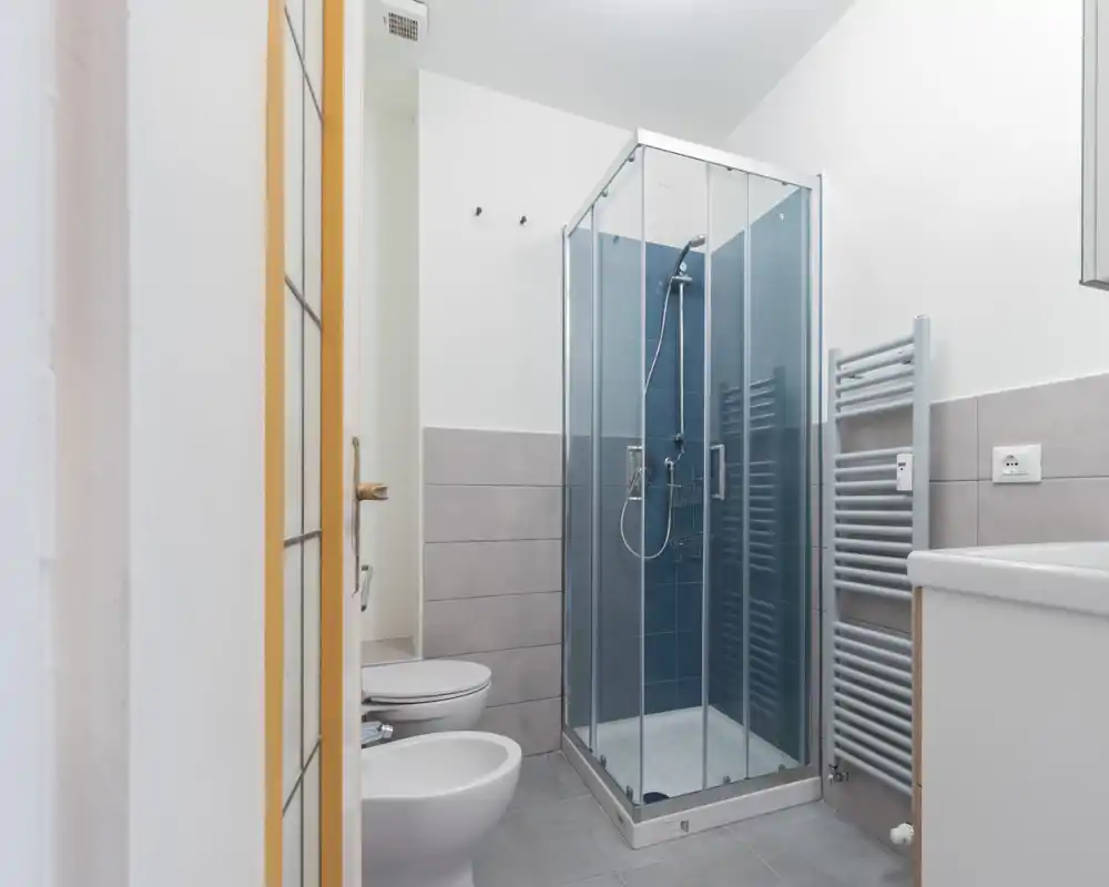 Baño moderno y compacto con ducha acristalada, bidé y wc, azulejos gris claro y toallero calefactable.