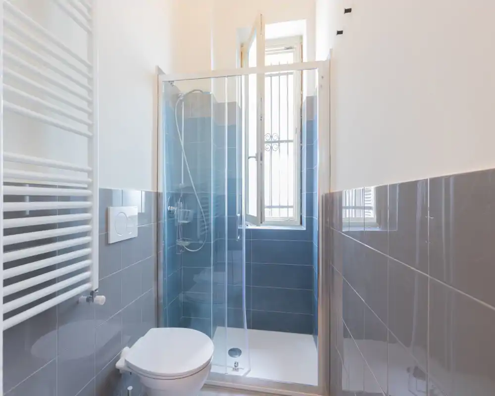 Baño compacto y moderno con ducha de esquina de cristal, inodoro suspendido y radiador toallero vertical; azulejos grises y ventana que proporcionan buena luz natural.