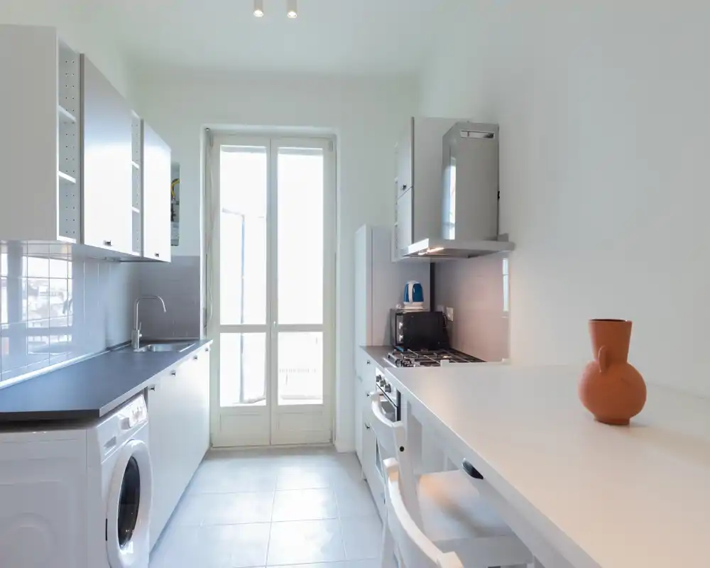 Cocina moderna y luminosa en galería con muebles blancos, encimera amplia, electrodomésticos integrados y una barra con un jarrón de terracota — ideal para un piso compacto.