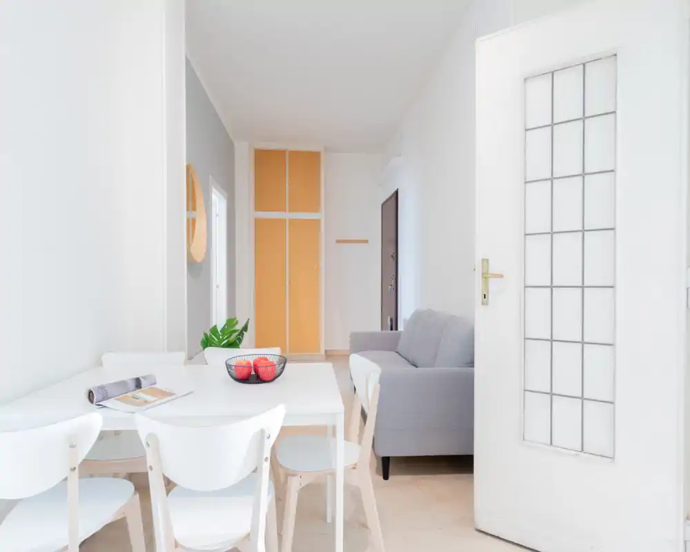 Zona de comedor luminosa y minimalista con mesa blanca y sillas a juego junto a una pequeña sala con sofá gris; estilo moderno y despejado ideal para anuncios.