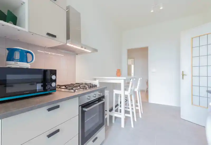 Cucina moderna e pulita con piano cottura a gas, forno incassato, microonde e un piccolo tavolo alto con sgabelli; pareti bianche luminose e arredi minimal creano un'atmosfera ariosa.