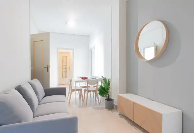 Soggiorno luminoso e moderno con un comodo divano grigio, mobile basso minimalista e uno specchio rotondo a parete; un piccolo tavolo da pranzo sullo sfondo crea un ambiente open-space arioso.