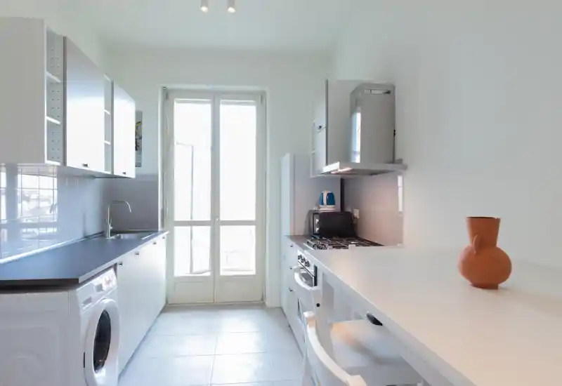 Cucina moderna e luminosa a corridoio con mobili bianchi, ampio piano di lavoro, elettrodomestici incassati e un piano colazione con una brocca in terracotta — perfetta per un appartamento compatto.